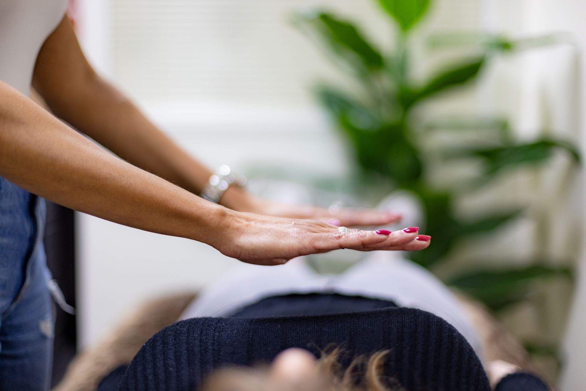 Person performing Reiki healing with hands hovering over another person lying down, plant in background.