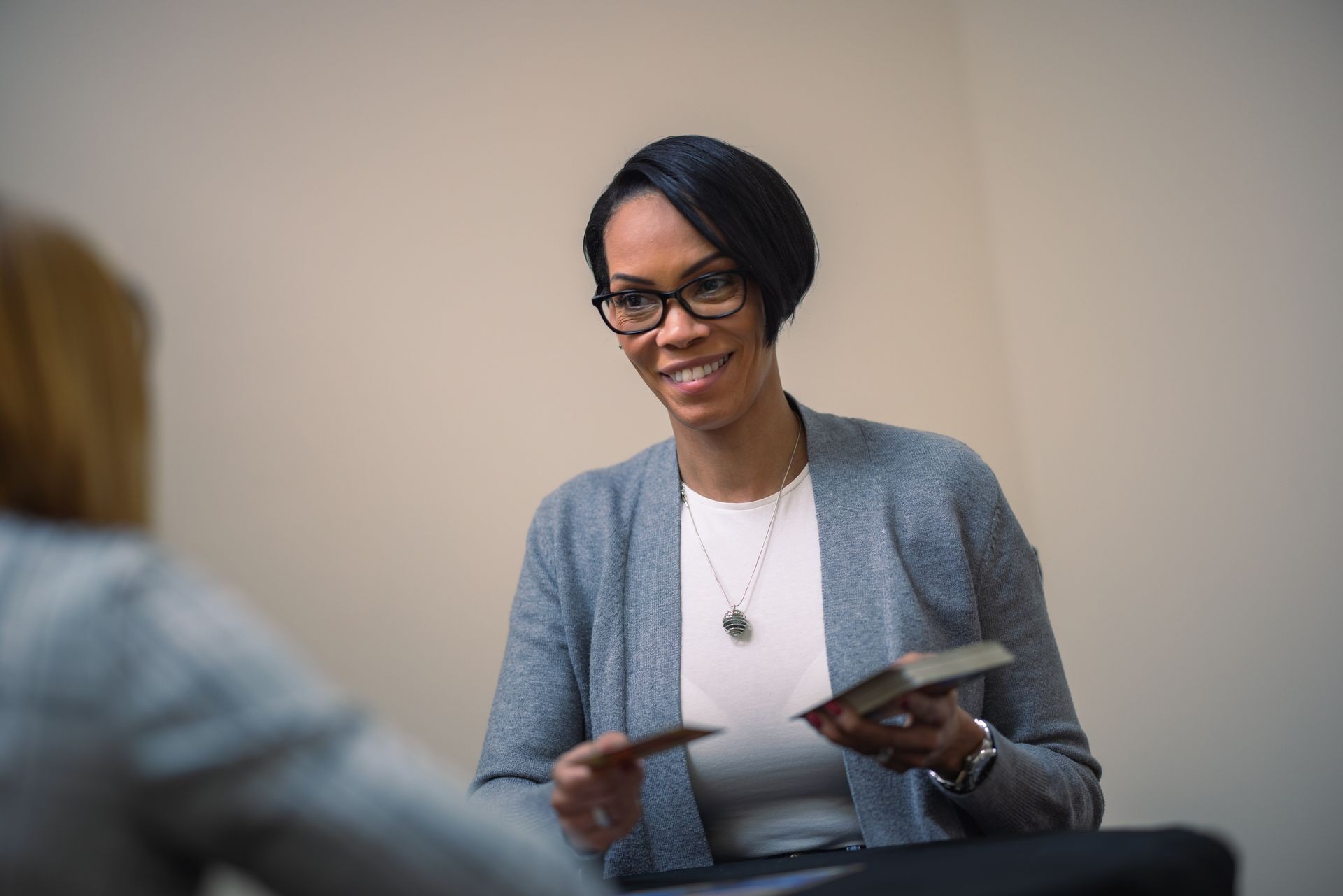 Woman wearing a gray sweater and holding cards, with another person facing her in a blurred background.